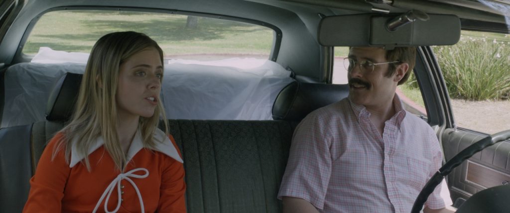 Two people sit in the front seats of a car, having a conversation. The woman wears an orange outfit, and the man in glasses wears a pink shirt. The background shows a grassy area.