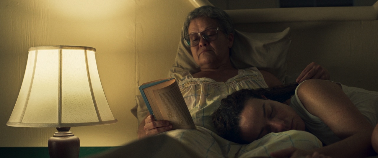 An older woman with glasses reads in bed beside a lamp while a younger person sleeps with their head resting on her arm.