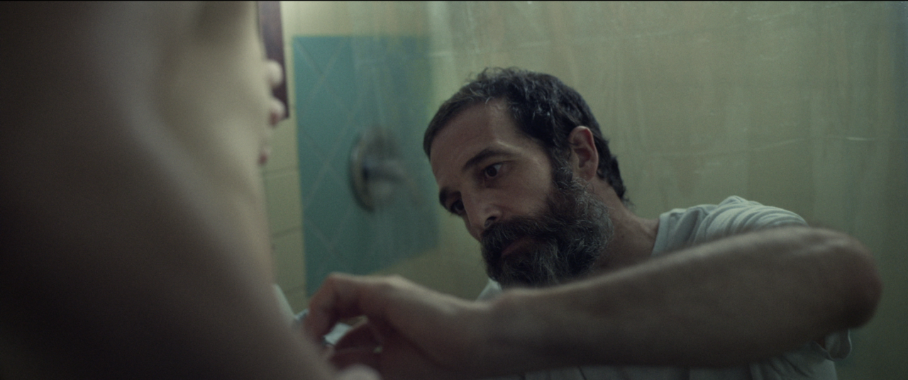 A bearded man looks intently at someone while adjusting something in a dimly lit bathroom.