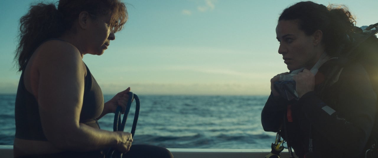 Two women sit on a boat at sea, one holding a rope and the other wearing scuba gear, facing each other with a calm ocean in the background.