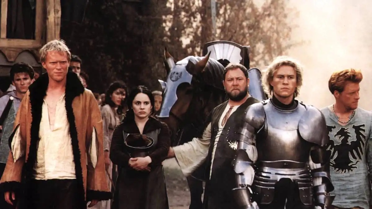 A group of people in medieval attire, including a knight in armor, stand together. A horse is visible in the background.