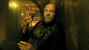 A man in tactical gear leans against a wall, holding a grenade. The background is dimly lit with visible wooden beams.