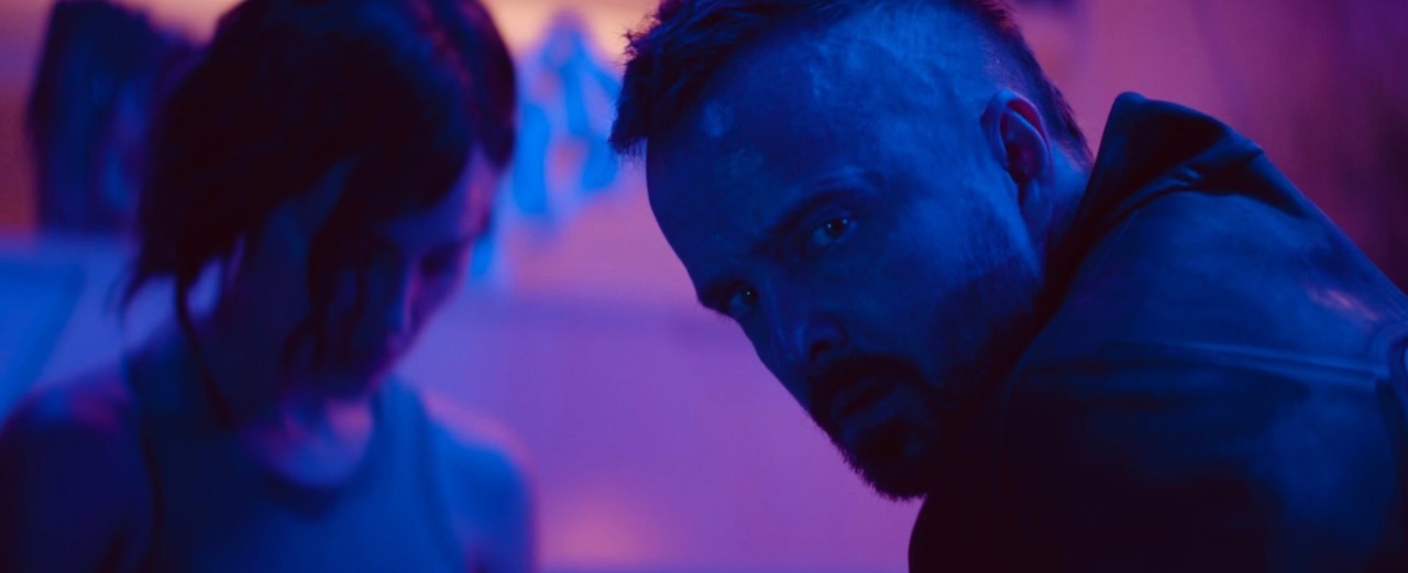 A person with short hair and a serious expression looks to the side in a dimly lit room with blue and purple lighting. Another person is blurred in the background.