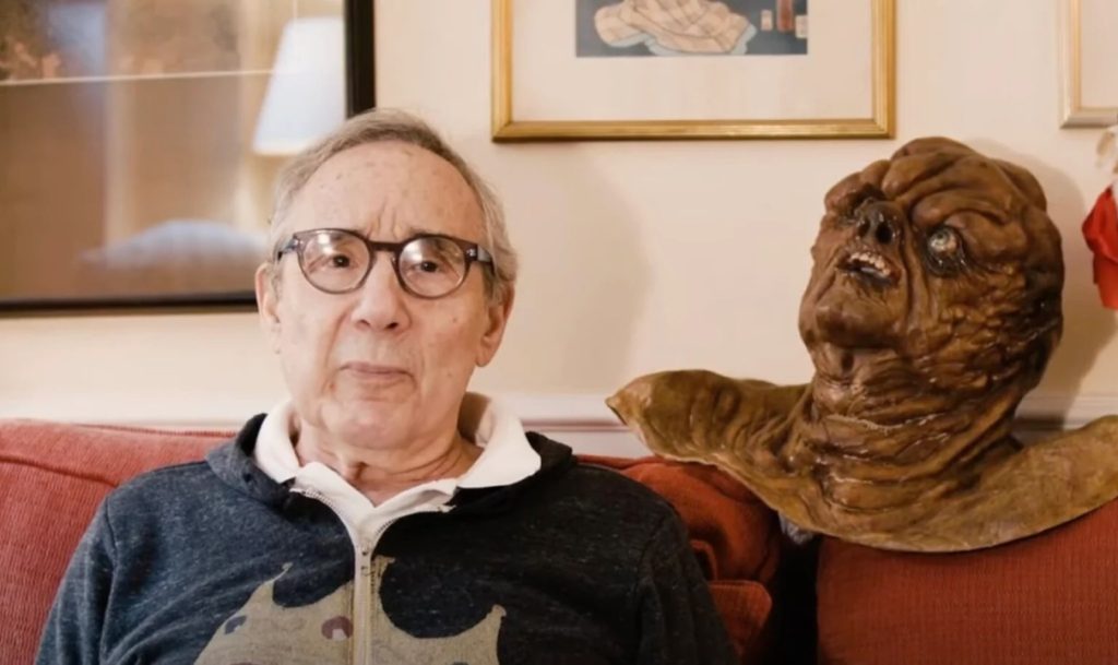An elderly man sitting on a red couch beside a grotesque creature sculpture. The background has framed artwork on the wall.