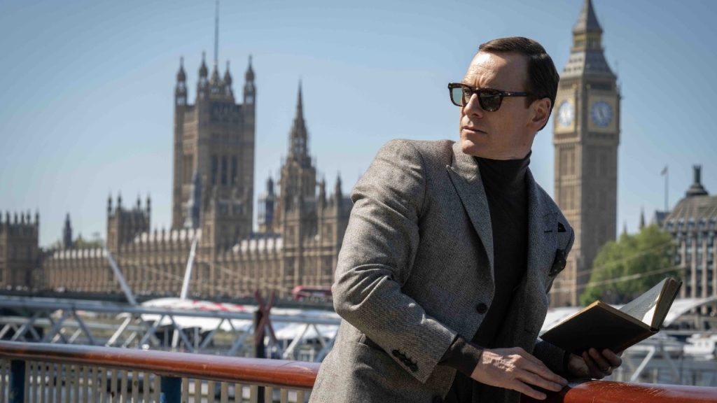 A person in sunglasses and a gray blazer leans on a railing while holding a notebook. The UK Parliament building and Big Ben are visible in the background.