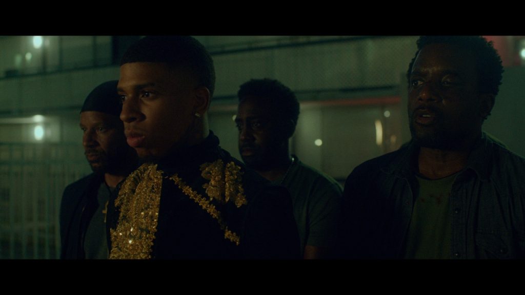 Four people stand together at night, one wearing a black and gold sequined top, with an urban background.