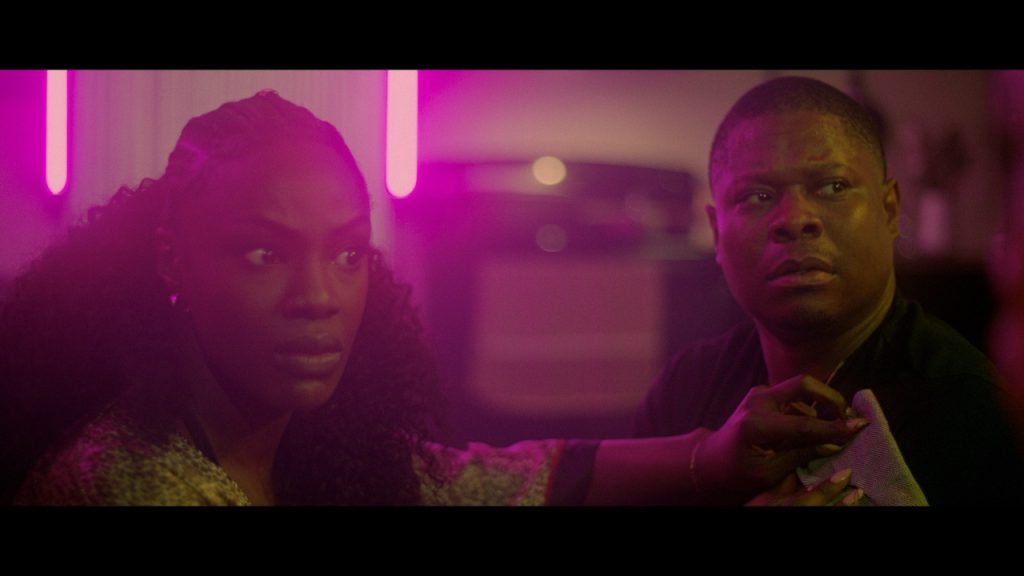 Two individuals look surprised in a dimly lit room with pink neon lighting. One is adjusting the other's collar.