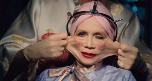 A person with a pink towel around their head and heavy makeup undergoes a cosmetic procedure, with their face skin being stretched by gloved hands using a metal frame.