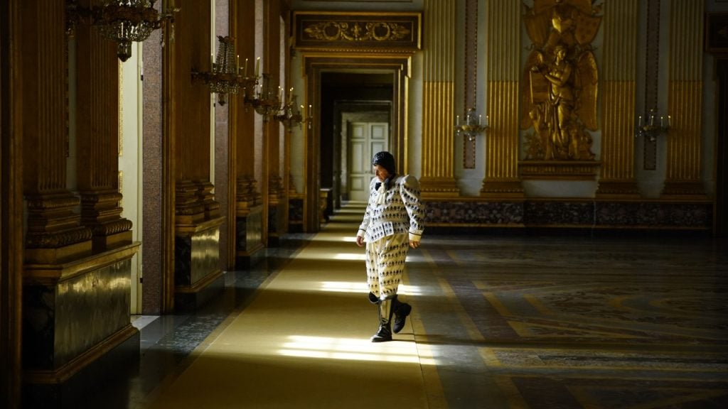A person in a patterned outfit walks through an ornate, sunlit hallway with decorative walls and chandeliers.