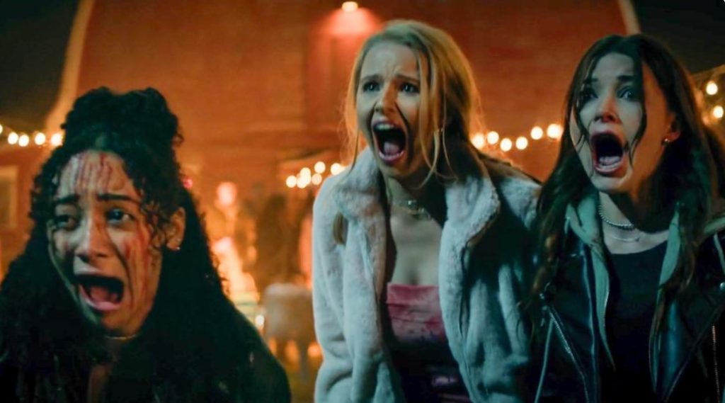 Three people are screaming, looking fearful. They are standing outside with lights and a building visible in the background.