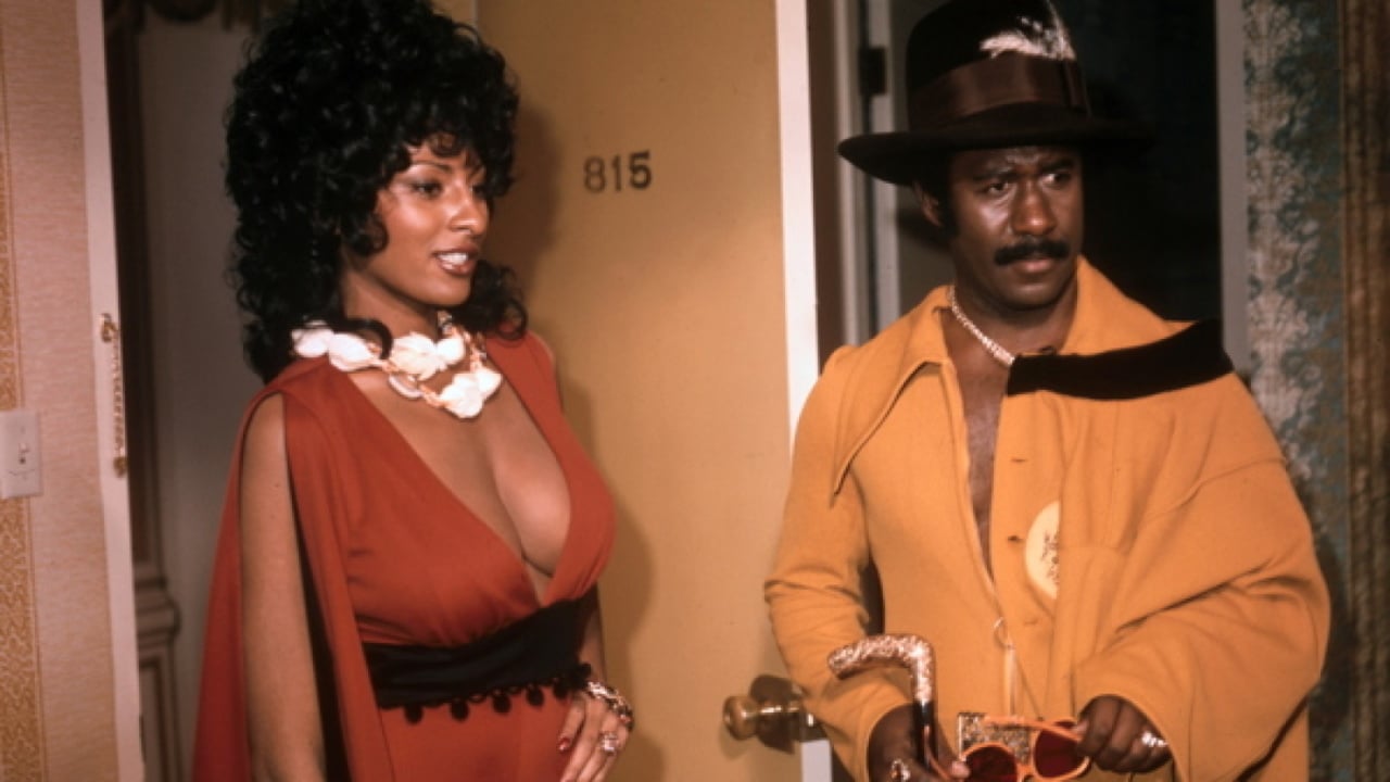 A woman in a red dress with a floral necklace stands beside a man in a yellow suit and black hat, in front of a door numbered 815.