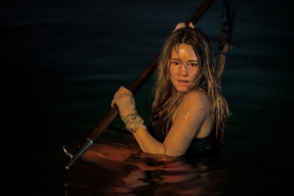 A person in a black outfit is waist-deep in water, holding a pole with both hands. Their wet hair is partially covering their face.