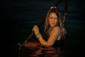 A person in a black outfit is waist-deep in water, holding a pole with both hands. Their wet hair is partially covering their face.