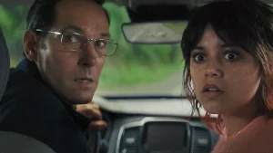 A man and a woman sit in a car, looking intently over their shoulders. The woman has short hair and a nose ring. The man wears glasses and a black shirt.