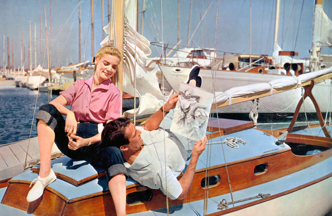 A man reclines on a sailboat, holding a sketch, while a woman sits nearby smiling. Docked boats are in the background.