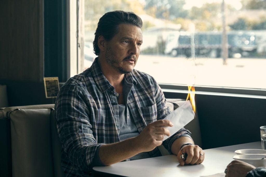 A man in a plaid shirt sits in a diner booth, holding a piece of burning paper. Sunlight streams through the window, illuminating the scene.