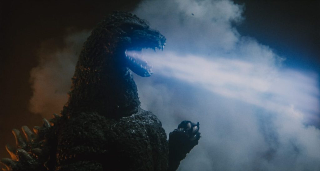 A giant dinosaur-like creature breathes blue atomic breath against a cloudy night sky.
