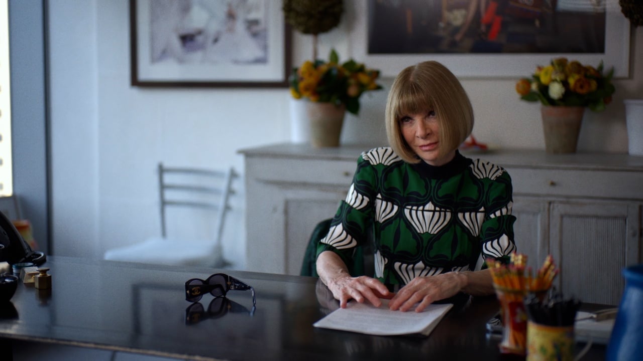 Person with a bob haircut sits at a desk with papers, sunglasses, and art supplies. Background includes framed art and flowers.
