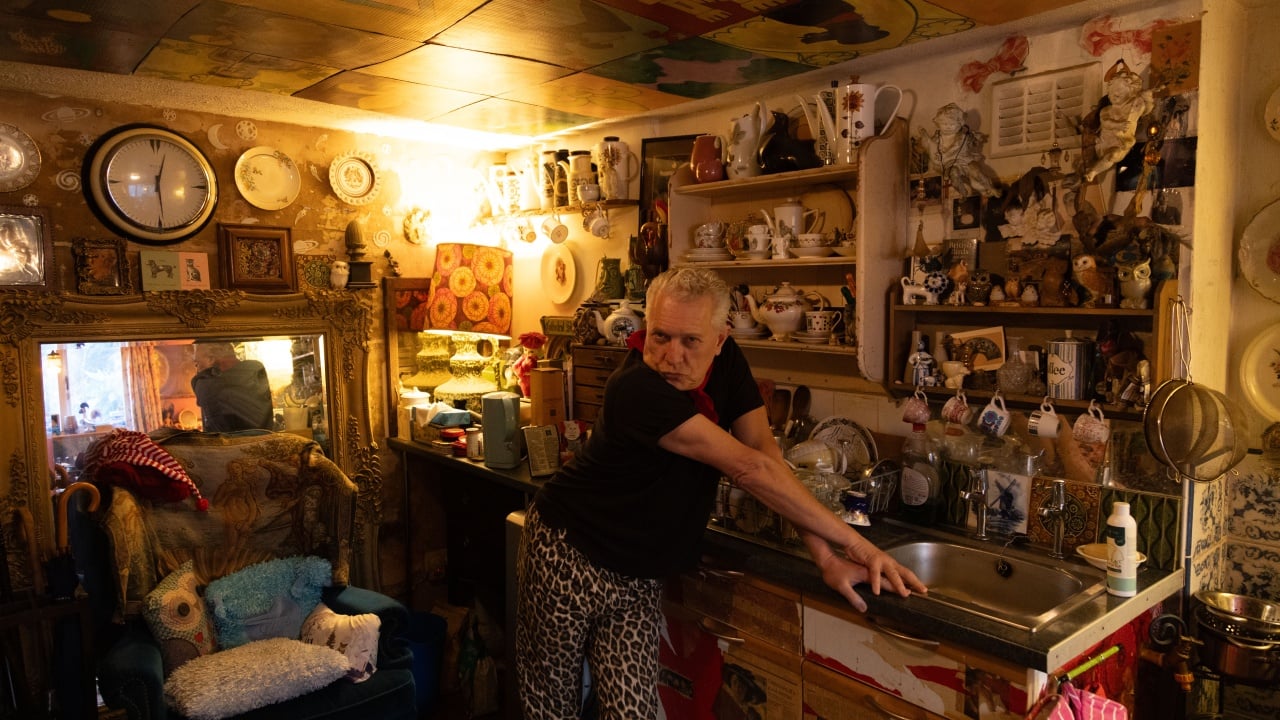 A person leans on a sink in a cluttered, eclectic room filled with various decorative items, mirrors, and paintings.
