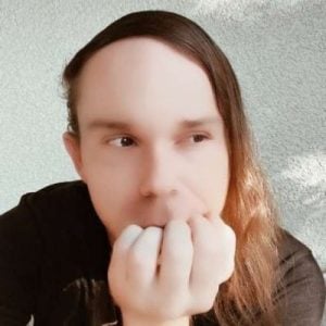Person with long hair and partially covering their face with their hand, wearing a black shirt, set against a textured, light-colored background.