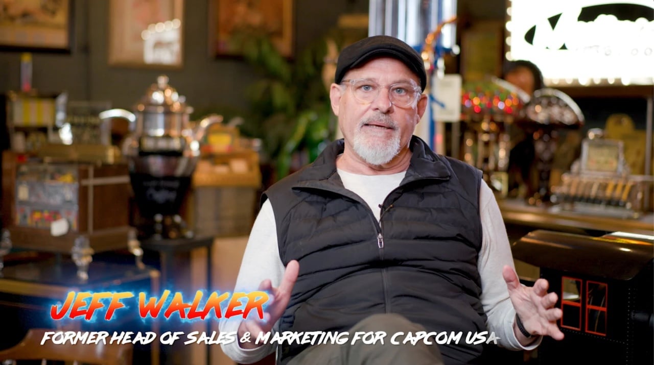 Seated man with glasses and cap, wearing a black vest and white shirt, gesturing with hands. Text overlay: "Jeff Walker, Former Head of Sales & Marketing for Capcom USA." Retro arcade setting.