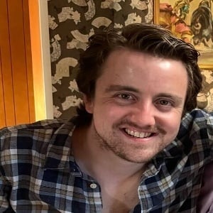 A person with brown hair and a beard wearing a blue plaid shirt, smiling in a room with patterned wallpaper and art.