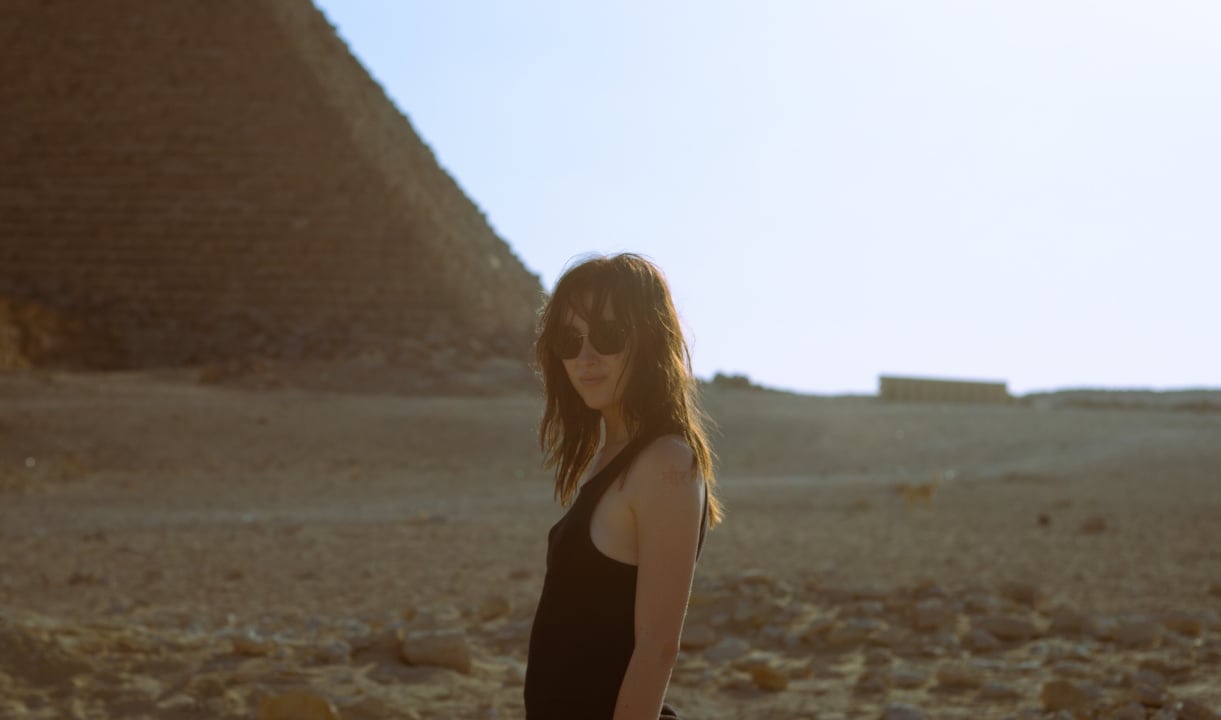 A person wearing sunglasses stands in front of a pyramid in a desert landscape.