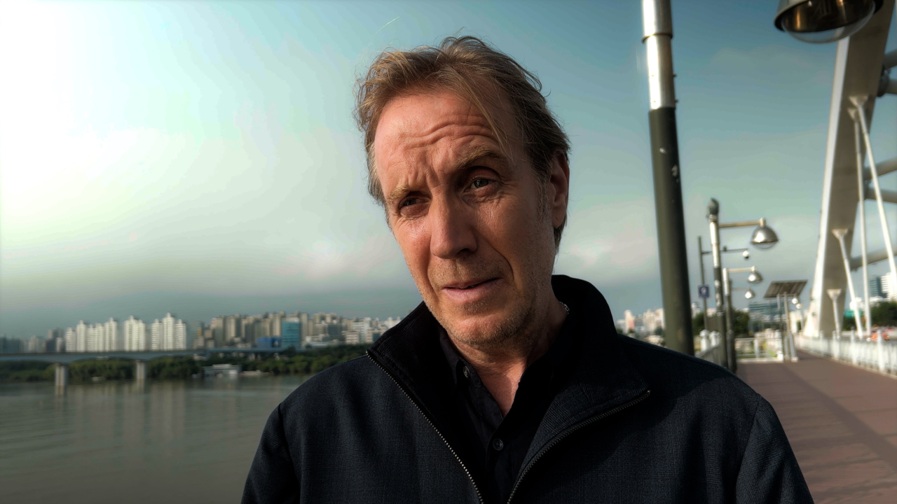 A person in a black jacket stands on a riverside walkway with a cityscape in the background.