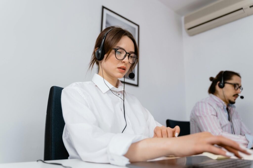 Two people in an office setting, wearing headsets. The person in the foreground types on a keyboard, while the other is seated in the background.