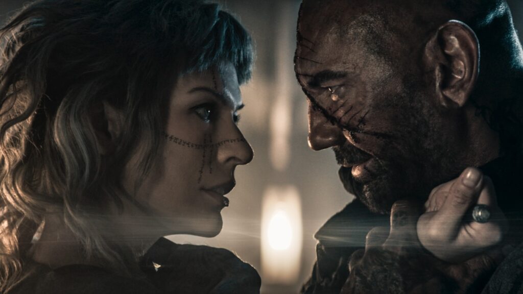 A woman and a man with facial tattoos and scars share an intense gaze, with the woman holding the man's collar. The lighting is dim and atmospheric.