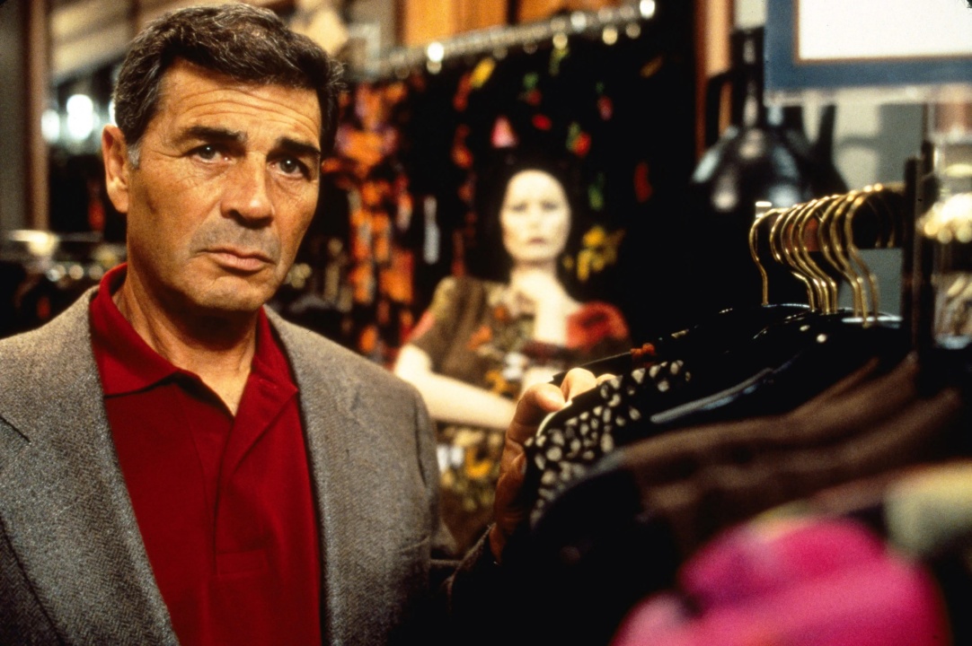 Man in a suit jacket browses clothing racks in a store, with colorful garments and a mannequin in the background.