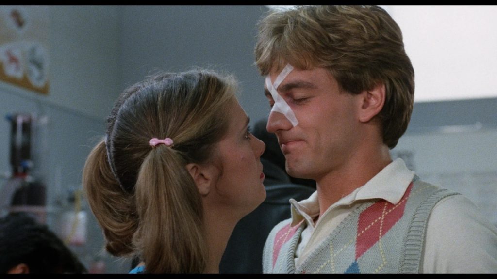 A woman with pigtails looks closely at a man with a bandage on his nose. The man wears a sweater vest and smiles.