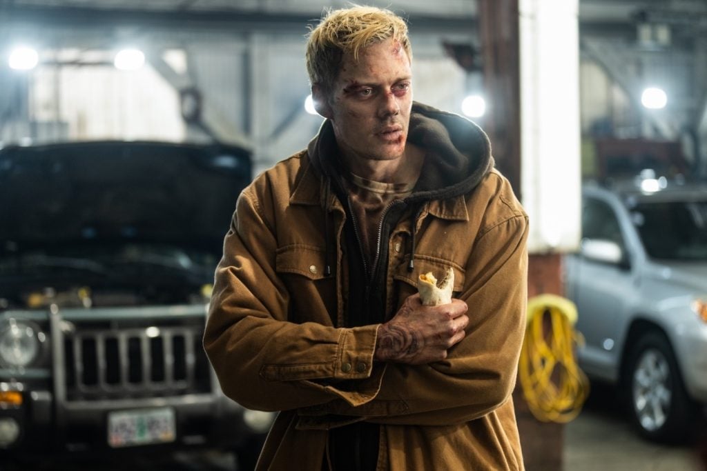 A man with bruises, wearing a brown jacket, holds a sandwich in a garage with cars in the background.