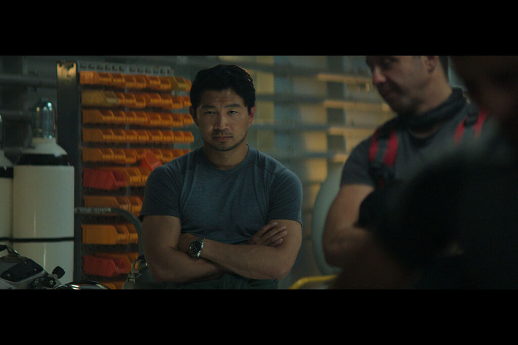 A person stands with arms crossed, wearing a gray t-shirt in a storage room with shelves of orange boxes.