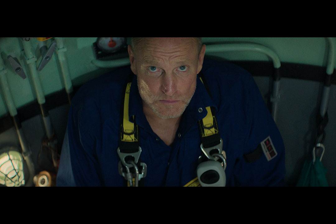 Person in a blue shirt with yellow harness looks up, sitting in a dimly lit, confined space with various equipment around.