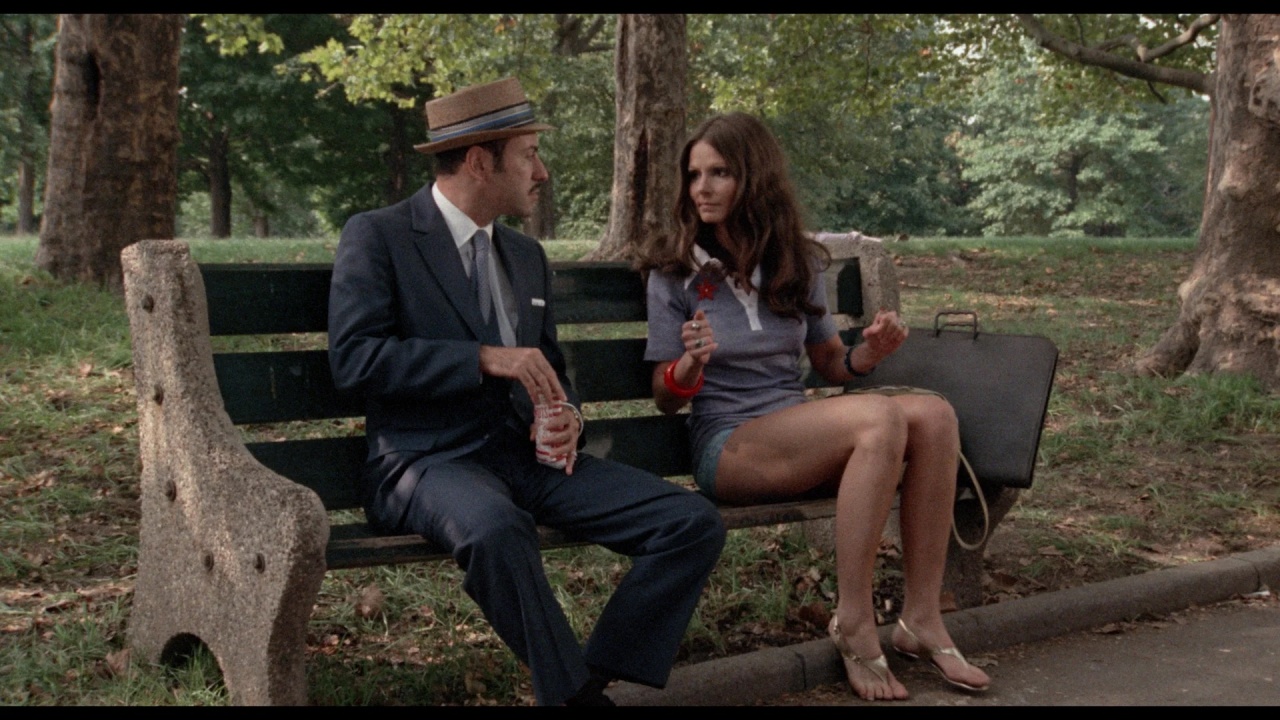 A man in a suit and hat sits on a park bench next to a barefoot woman holding a rose and sunglasses. A briefcase is placed beside her. Trees are visible in the background.