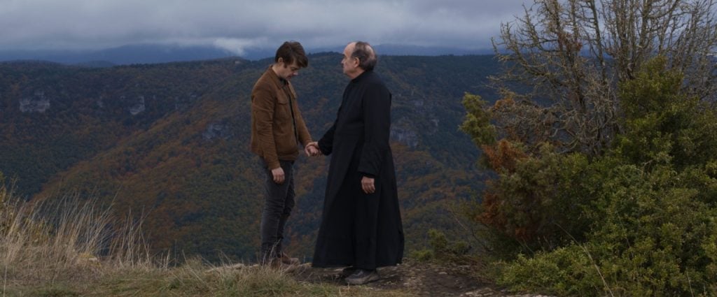 Two people stand on a scenic hillside; one in a brown jacket, the other in black clergy attire, holding hands and conversing with a hilly landscape in the background.