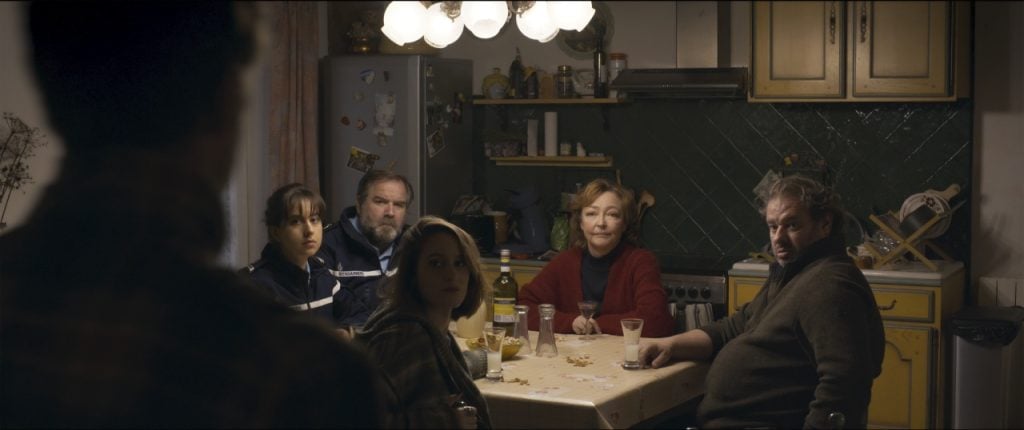 Six people sit around a table in a kitchen with dim lighting. They appear focused on someone or something out of the frame.
