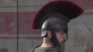 A person wearing an ancient Roman-style helmet with a red crest stands in profile against a stone wall with faded red and beige designs.