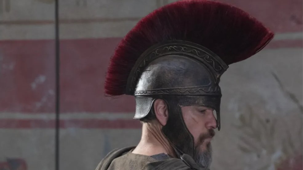 A person wearing an ancient Roman-style helmet with a red crest stands in profile against a stone wall with faded red and beige designs.