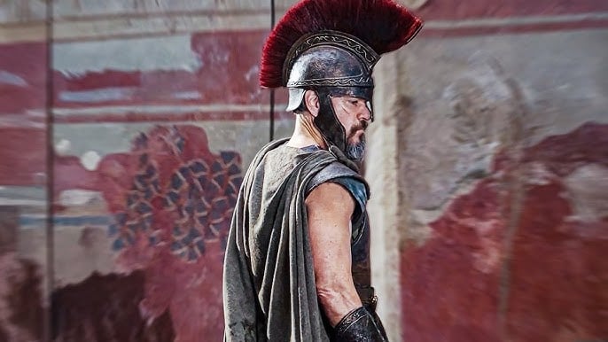 A person in ancient warrior attire, including a plumed helmet and a cloak, stands against a painted wall background.