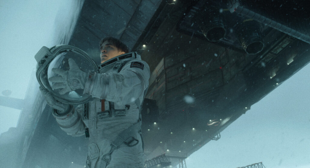 An astronaut in a white spacesuit holds a helmet, standing under a large spacecraft in a snowy environment.