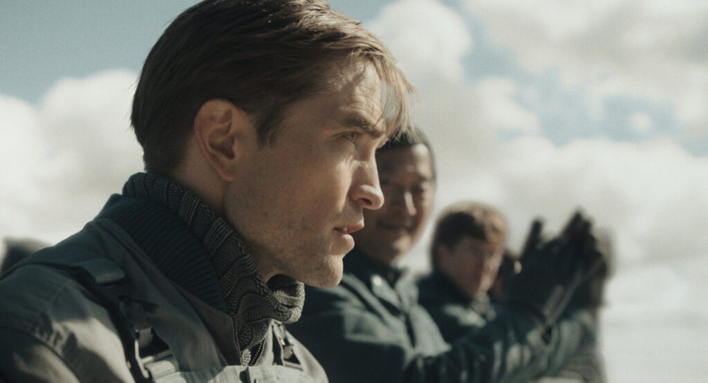 Three people wearing outdoor jackets stand in a line under a cloudy sky, with one person in focus gazing into the distance.