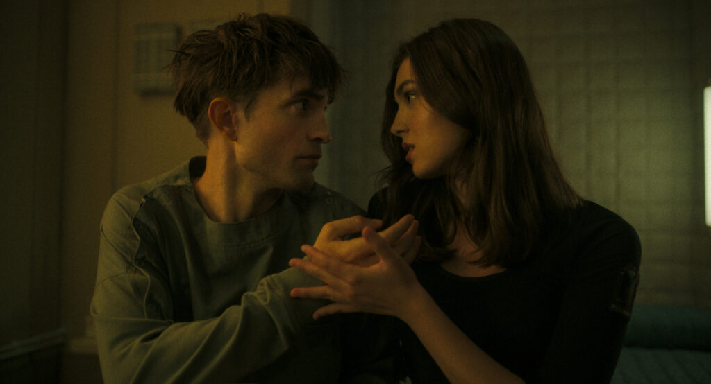 A man and a woman sit closely, gazing at each other intently, in a dimly lit room. The man wears a light-colored shirt, while the woman is in a dark top, and they hold hands.