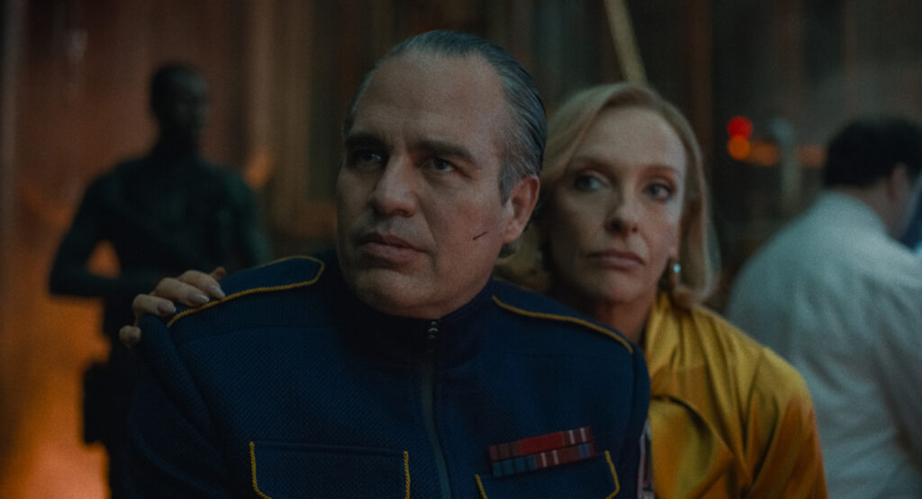 A man in a blue uniform with medals and a scar on his cheek sits with a woman in a yellow top, who has her hand on his shoulder. They appear to be observing something off camera.