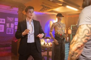 A man in a suit stands in a fighting stance in a dimly lit room with a mannequin, wall art, and neon lights in the background.