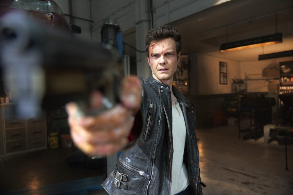 Person in a leather jacket points a gun, appearing intense, inside a dimly lit garage.