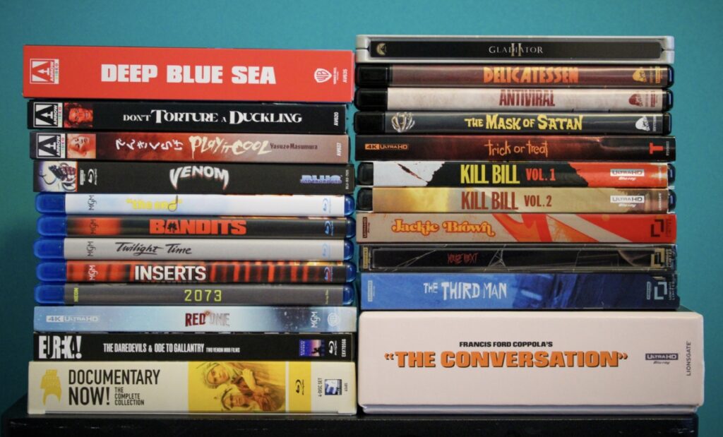 A collection of DVD and Blu-ray cases are stacked against a turquoise background, featuring titles like "Deep Blue Sea," "Kill Bill Vol. 1," and "The Conversation.