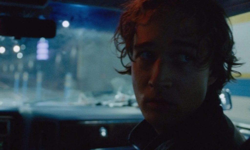 Young person with curly hair sits in the front seat of a car at night, looking toward the back. The background is dimly lit with reflections on the windshield.