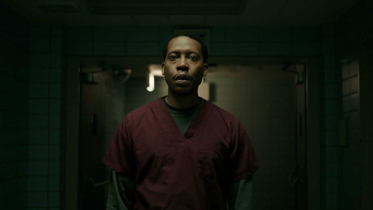 A person in maroon scrubs stands in a dimly lit hallway, looking forward. The background features tiled walls and partially open doors.